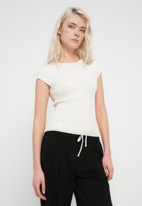 Top de canalé color crema con pequeñas mangas tipo capa, silueta ajustada y cuello redondo. Combinado con pantalones negros con cintura de cordón.