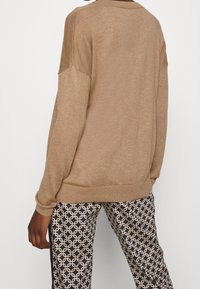 Beige gebreide trui met een ronde hals en doorgezakte schouders, gecombineerd met zwart-witte broek met een patroon van in elkaar grijpende vormen.