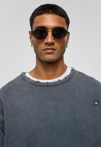 Ronde zwarte zonnebril, grijze trui met versleten halslijn en stof, gelaagd over een wit shirt. Soepele textuur, casual pasvorm.