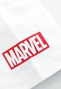 Vit bomullströja med en framträdande röd rektangulär logotyp med texten "MARVEL" i fet stil i vitt. Slät textur.