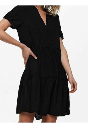 Vrouw die een zwarte jurk met korte mouwen en lagen draagt, met een V-hals, poserend met één hand op haar heup en de andere ontspannen langs haar zijde.