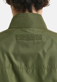 Giacca verde, leggera, con una texture liscia. Presenta un colletto strutturato e un logo discreto ricamato sul retro.