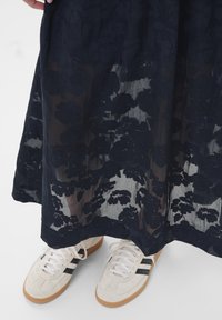 Jupe longue marine à motif floral avec une superposition transparente, associée à des chaussures de sport blanches avec des rayures noires et une semelle gomme.