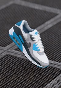 Nike Air Max 90 sneaker featuring a grey, white, and blue color scheme, mesh and synthetic materials, distinct air cushioning, and lace closure.