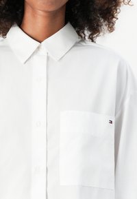 Camisa blanca de botones con collar clásico, un bolsillo en el pecho y un pequeño logotipo de marca en rojo, blanco y azul en el bolsillo.
