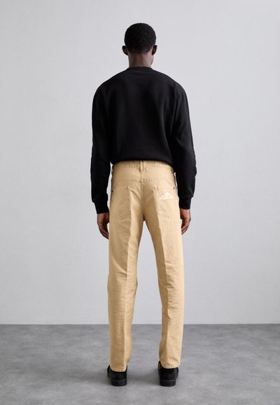 Sweatshirt noir, pantalon en coton beige avec poches arrière, design simple et détail du logo blanc ; chaussures noires ; vue de dos.