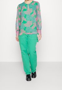 Blusa floreale a maniche lunghe verde, con motivi rosa e blu; abbinata a pantaloni verdi dalla vestibilità ampia e calzature nere.