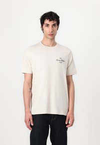 Beige cotton T-shirt with short sleeves, crew neck, and a small black logo design on the left side. Worn with dark jeans.