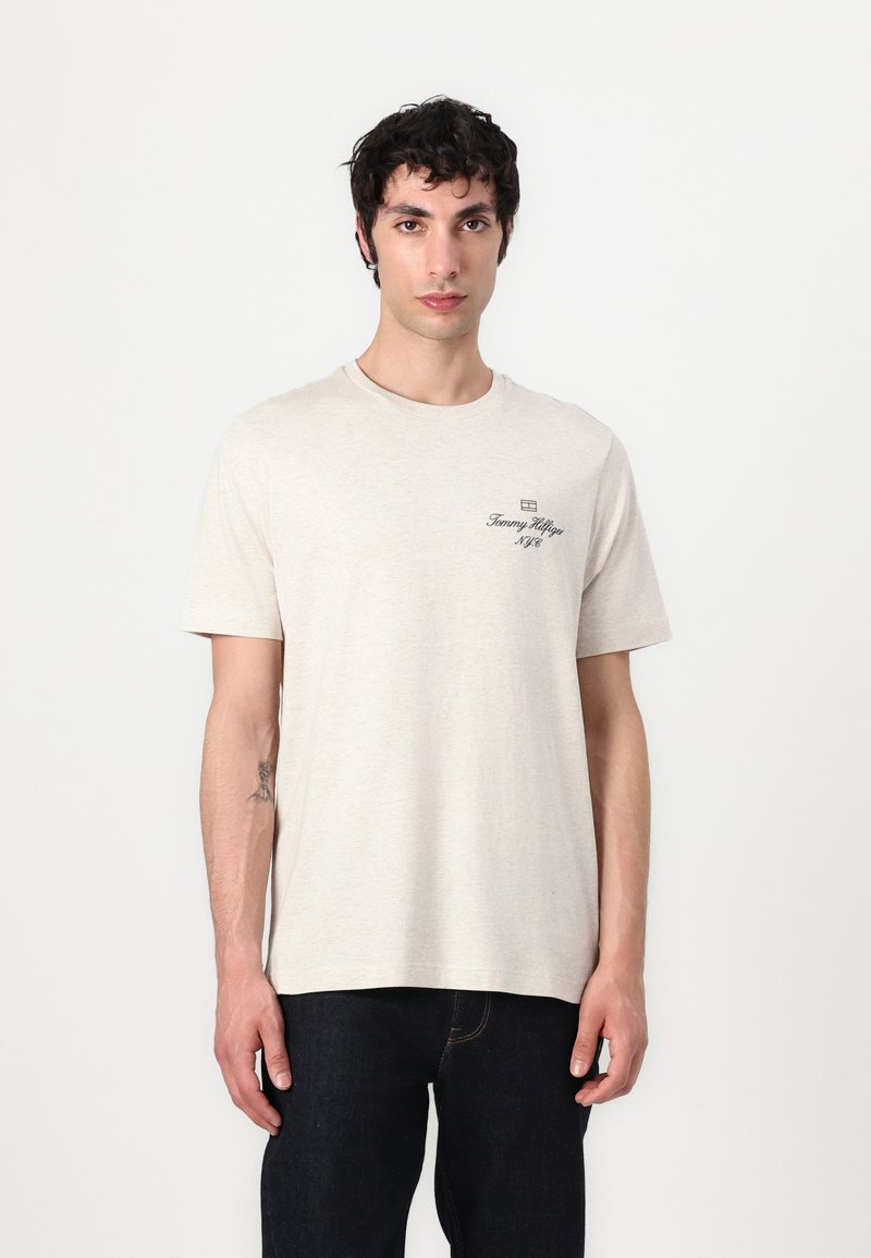 Beige bomulds T-shirt med korte ærmer, rund hals og et lille sort logo-design på venstre side. Båret med mørke jeans.