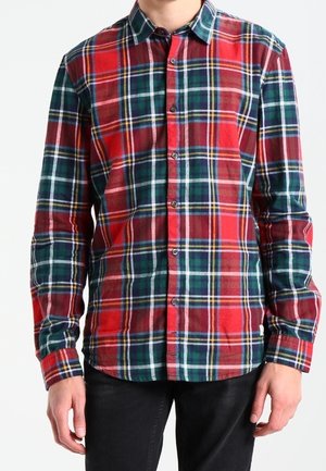 Mężczyzna ubrany w czerwoną, zieloną i granatową koszulę w kratę na guziki z długimi rękawami oraz czarne spodnie, stojący z rozluźnionymi ramionami.