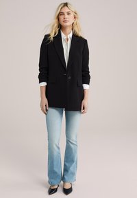 Černý přizpůsobený blazer s klopami, s jedním knoflíkem a kapsami, zkombinovaný s bílou košilí a světle modrými rozšířenými džíny.