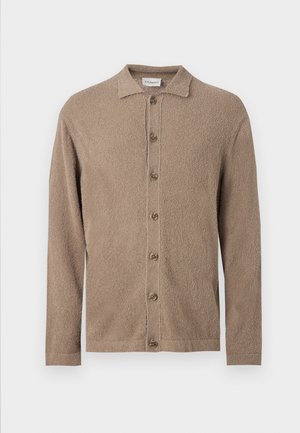 Jeune homme portant une chemise beige, un cardigan marron boutonné, un pantalon ample foncé et des chaussures habillées noires, debout les mains dans les poches.