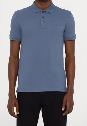 Uomo che indossa una polo blu a maniche corte con tre bottoni e pantaloni neri, in piedi davanti a uno sfondo bianco uniforme.