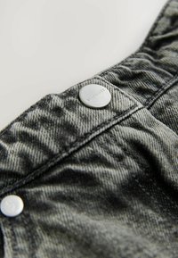 Sezione di abbigliamento in denim caratterizzata da un tessuto grigio scuro, cuciture in vista e un bottone metallico decorato con il nome del marchio inciso, AGUASANTS.