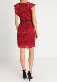 Robe rouge en dentelle avec des manches courtes évasées, une bordure noire et une taille cintrée. L'ourlet présente un bord festonné. Portée avec des chaussures à talons hauts noires.