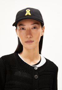 Schwarze Baseballmütze mit gelbem gesticktem Logo, kombiniert mit einem schwarzen Strickcardigan mit Knopfdetails und einer weißen Innenschicht.