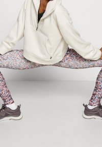 Biały polarowy sweter z kieszenią z przodu, połączony z wzorzystymi legginsami w kolorze różowym, niebieskim i fioletowym. Szare buty sportowe i białe skarpetki.
