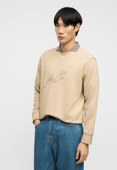 Νέος άνδρας που φοράει μπεζ φούτερ με κείμενο σε γραφή πάνω από πουκάμισο με γιακά, με τα χέρια στις τσέπες και τζιν μπλε, στέκεται μπροστά σε απλό φόντο.