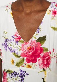 Blommig klänning med vit bakgrund och starka rosa pioner, lila blommor och gröna blad. V-halsdesign med mjuk textur.