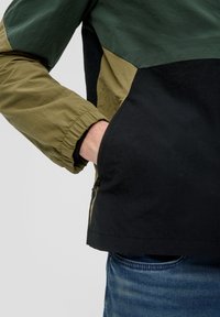 Kleurgeblokte jas met donkere groene, olijfkleurige en zwarte panelen. Lichtgewicht stof met elastische manchetten en een ritszak aan de zijkant.