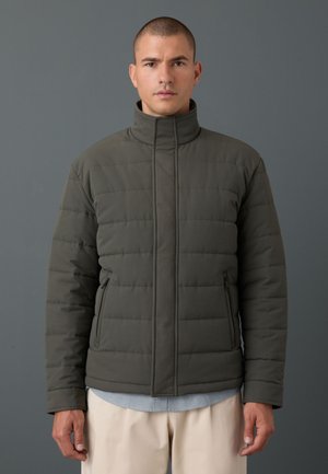 Mann trägt eine dunkelgrüne gesteppte Jacke mit hohem Kragen, beige Hose und ein hellblaues Hemd darunter, steht vor einem schlichten grauen Hintergrund.