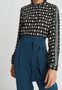 Blusa nera con motivi geometrici; maniche lunghe; abbinata a pantaloni teal a vita alta con un dettaglio di cintura annodata.