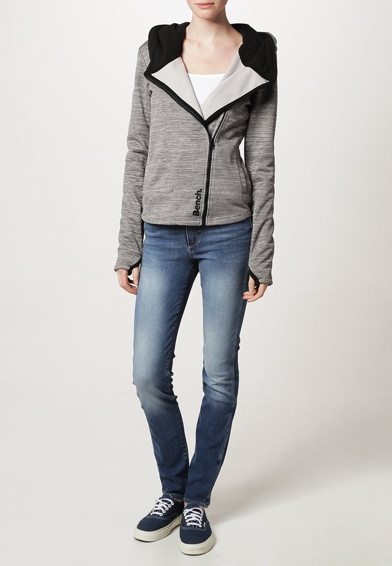 Femme portant un sweat à capuche gris zippé avec une capuche noire, un jean skinny bleu, un haut blanc et des baskets bleu marine à lacets blancs.