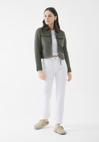 Olivgrüne Lederjacke im Cropped-Stil mit zwei Brusttaschen, kombiniert mit weißen Jeans aus Denim und beigen Slippern. Glatte Textur, minimalistisches Design.