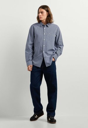 Jonge man met schouderlang bruin haar, die een blauw geruit overhemd met lange mouwen, donkerblauwe spijkerbroek en zwarte sneakers draagt, staand tegen een eenvoudige muur.