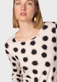 Maglia a maniche lunghe in crema con grandi pois neri. Presenta un colletto rotondo e una morbida texture del tessuto. Modella che indossa orecchini geometrici dorati.