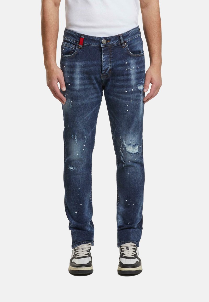 Hombre vestido con jeans oscuros desgastados y manchas de pintura blanca, y zapatillas en blanco y negro, de pie contra un fondo liso.