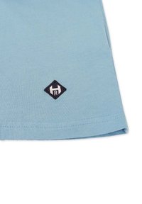 Vaalean sininen puuvillapaita, jossa on pyöreä helma. Paidan alakulmassa on musta timantin muotoinen logo, jossa on brodeerattuna "H" ja "R".