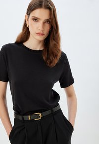 Sort T-shirt lavet af blødt stof, med rund hals og korte ærmer, parret med højtaljede bukser og et grønt bælte med guldlukning.
