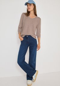 Maglione beige a V con texture, jeans blu a gamba larga, cappellino blu chiaro, sneakers beige e bianche. Posizione in piedi con le mani nelle tasche.