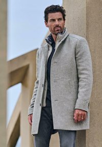 Hellgrauer Wollmischmantel mit strukturiertem Kragen, texturiertem Stoff und Knopfverschluss, getragen über einem dunkelblauen Hemd und grauen Jeans.