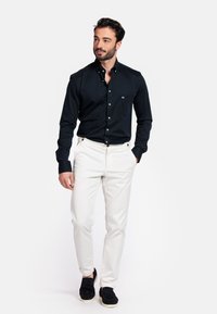 Marineblauw overhemd met knopen, voorzien van een linker borstzak, gecombineerd met witte broeken en zwarte suède schoenen, met een gladde stoftextuur.