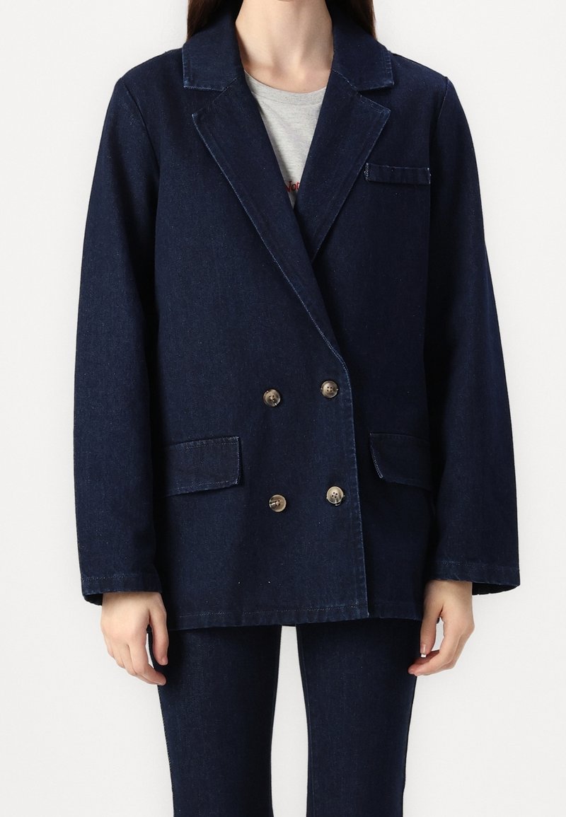 Veste blazer croisée en denim foncé avec larges revers, deux poches avant, boutons dorés et coupe ample. Tissu texturé avec un léger éclat.