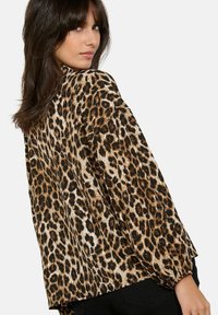 Mujer con cabello largo y oscuro, vestida con una blusa suelta de estampado de leopardo con mangas abullonadas, girando ligeramente para mostrar la parte trasera de la blusa.
