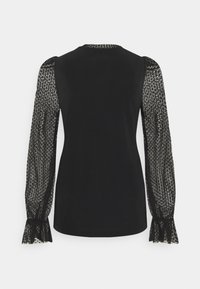 Haut noir à manches longues avec corps en tissu uni et manches en dentelle transparente à motifs se terminant par des poignets à volants, vu de dos.
