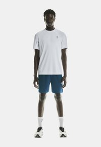 T-shirt blanc à manches courtes avec un logo minimal, associé à un short de sport bleu. Le modèle porte également des chaussettes blanches et des chaussures noires.