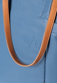 Borsa a tracolla blu con superficie liscia, manico in pelle marrone e logo impresso. Presenta un design minimal e una forma strutturata.