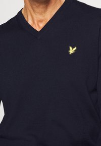 Pull col en V bleu marine en tissu tricoté, avec un petit logo d'oiseau jaune sur la poitrine gauche. Texture lisse, coupe régulière.