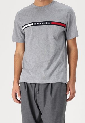 Homme portant un t-shirt gris à manches courtes avec une bande horizontale du logo Tommy Hilfiger sur la poitrine et un pantalon gris foncé.