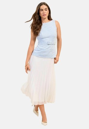 Kobieta z długimi brązowymi włosami, ubrana w niebieski bezrękawnik z paskiem i białą plisowaną spódnicę midi, chodząca w beżowych szpilkach ze złotymi końcówkami.