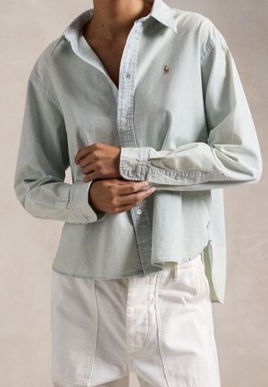 Persona ajustando el puño de una camisa verde claro con botones, combinada con pantalones cortos blancos, de pie frente a un fondo liso.