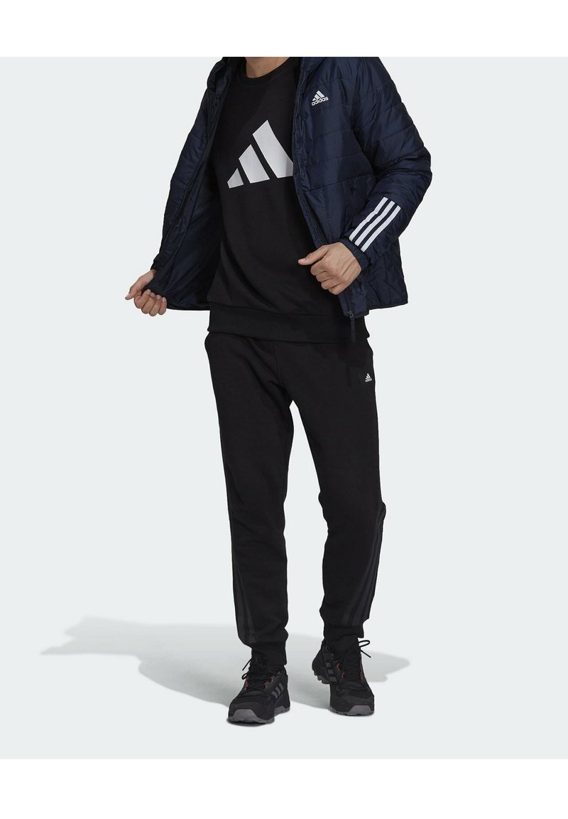 Námořnická péřová bunda, černá mikina s velkým bílým logem, černé tepláky, černé sportovní boty s červenými detaily.