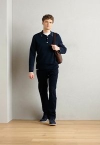 Marineblå polo med hvite detaljer, mørke bukser og sneakers. Brun skulderveske. Stående på et tregulv mot en grå vegg.