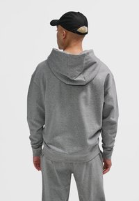 Osoba znajdująca się tyłem, ubrana w szary hoodie i pasujące dresy, z czapką na głowie, spod której widać rozjaśnione włosy.