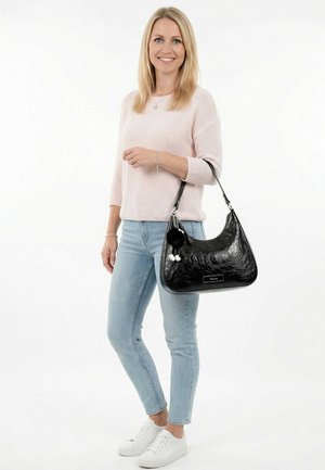 Lachende vrouw die een lichtroze trui, blauwe spijkerbroek en witte sneakers draagt, en een zwarte leren handtas vasthoudt met decoratieve bedels aan de band.