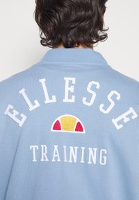Jasnoniebieski sweter z prążkowanym kołnierzem. Posiada biały haftowany napis "ELLESSE" oraz "TRAINING", a także wielokolorowy wzór logo.